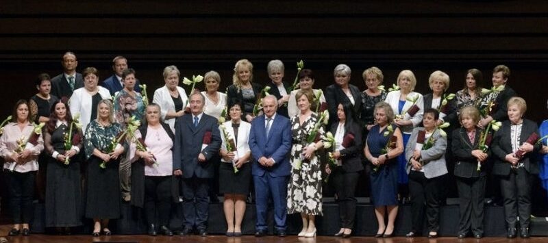 Díjazott üzletvezetők csoportképe egy színpadon, virágot tartva, a Coop jubileumi ünnepségén.