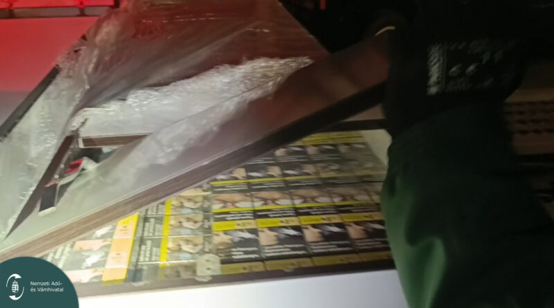 Egy pénzügyőr felfeszíti a bútorlapot, alatta jól láthatók a sárga-fekete csomagolású cigarettás dobozok.