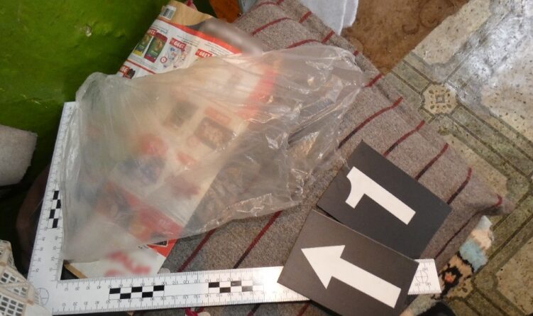 Átlátszó nejlonzacskóban lévő papírok és tárgyak egy szőnyegen, mellette mérőskála és irányt jelző bizonyítékjelölő táblák.