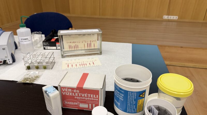 Laboratóriumi asztalon elhelyezett vér- és vizeletvételi egységdoboz, mintatároló edények és toxikológiai vizsgálati eszközök egy szakmai bemutatón.