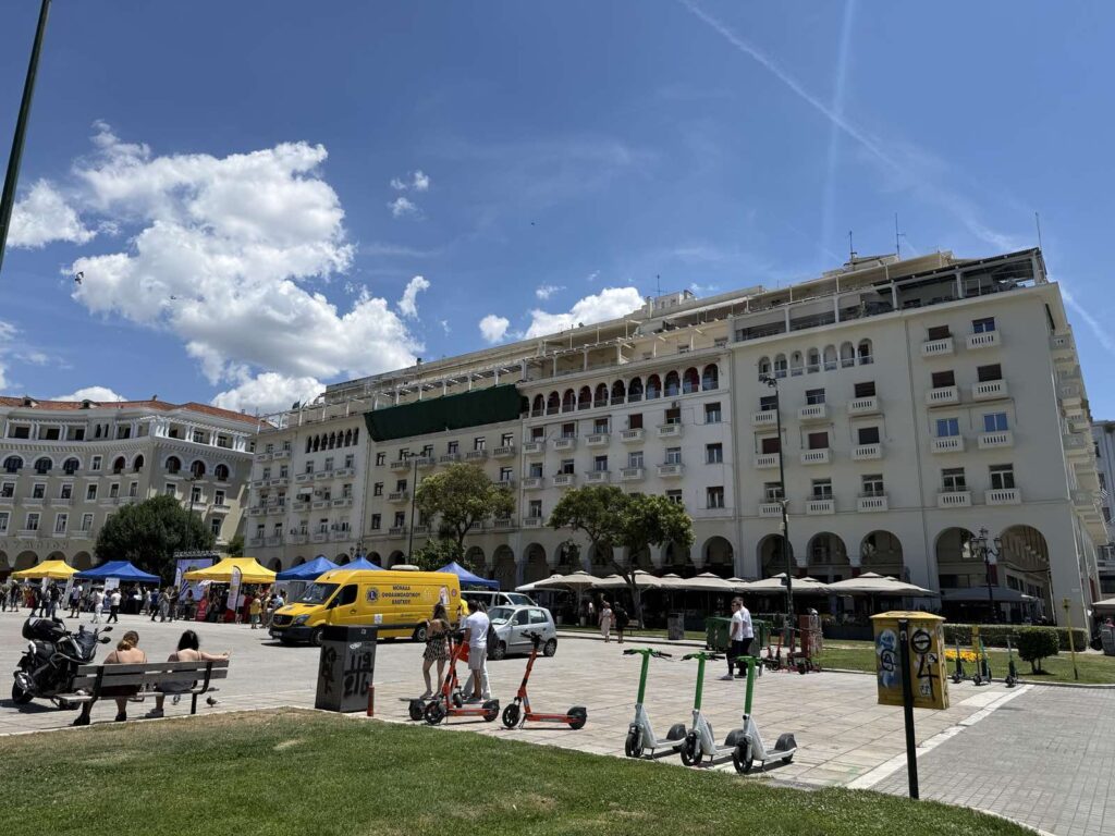 Thessaloniki főtere napsütésben, sétáló emberekkel és történelmi épületekkel