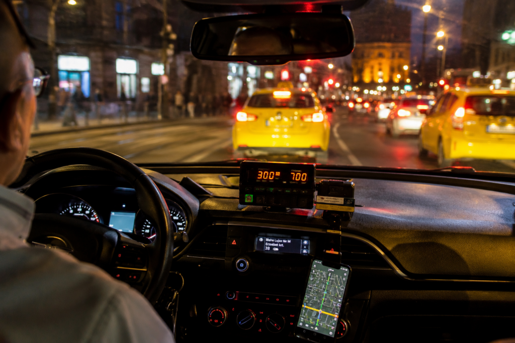 Budapesti taxi belső tere esti forgalomban, taxiórával és sárga taxikkal a városi fények között.