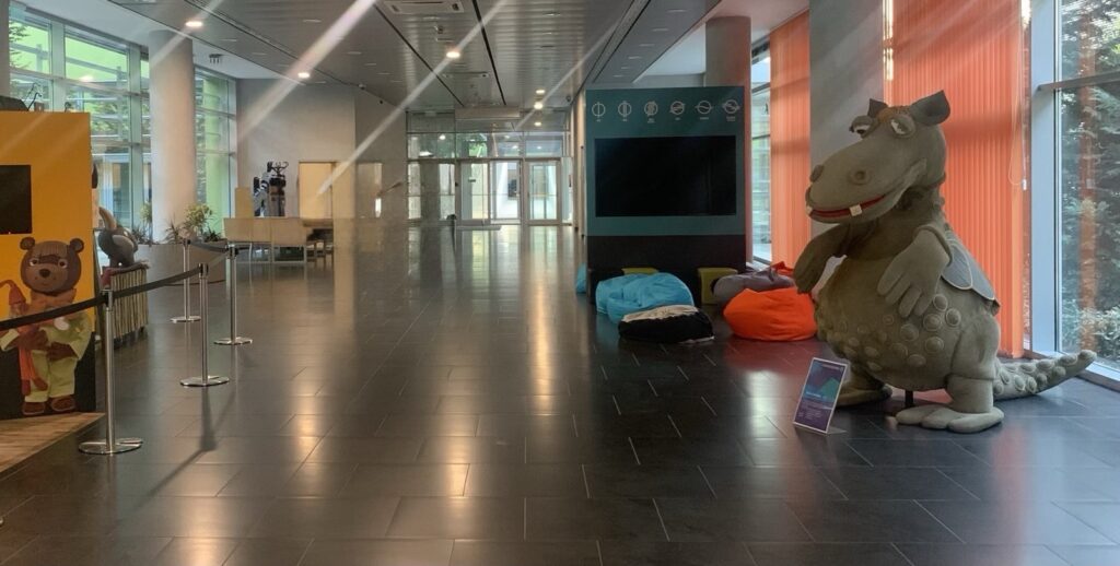 A TV Maci és Süsü, a sárkány figurái egy modern közmédiai épület előcsarnokában, interaktív gyereksarok közelében.