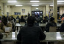 Menekültek ülnek egy nyugat-európai befogadóközpont étkezőjében, miközben a háttérben csendesen zajlik a tábor mindennapi élete.