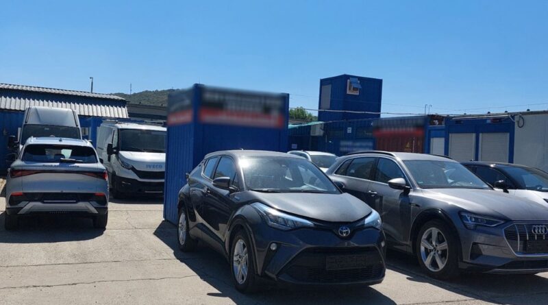Toyota és Audi személyautók, valamint haszonjárművek a kék konténerépületek előtti autókereskedői udvaron.