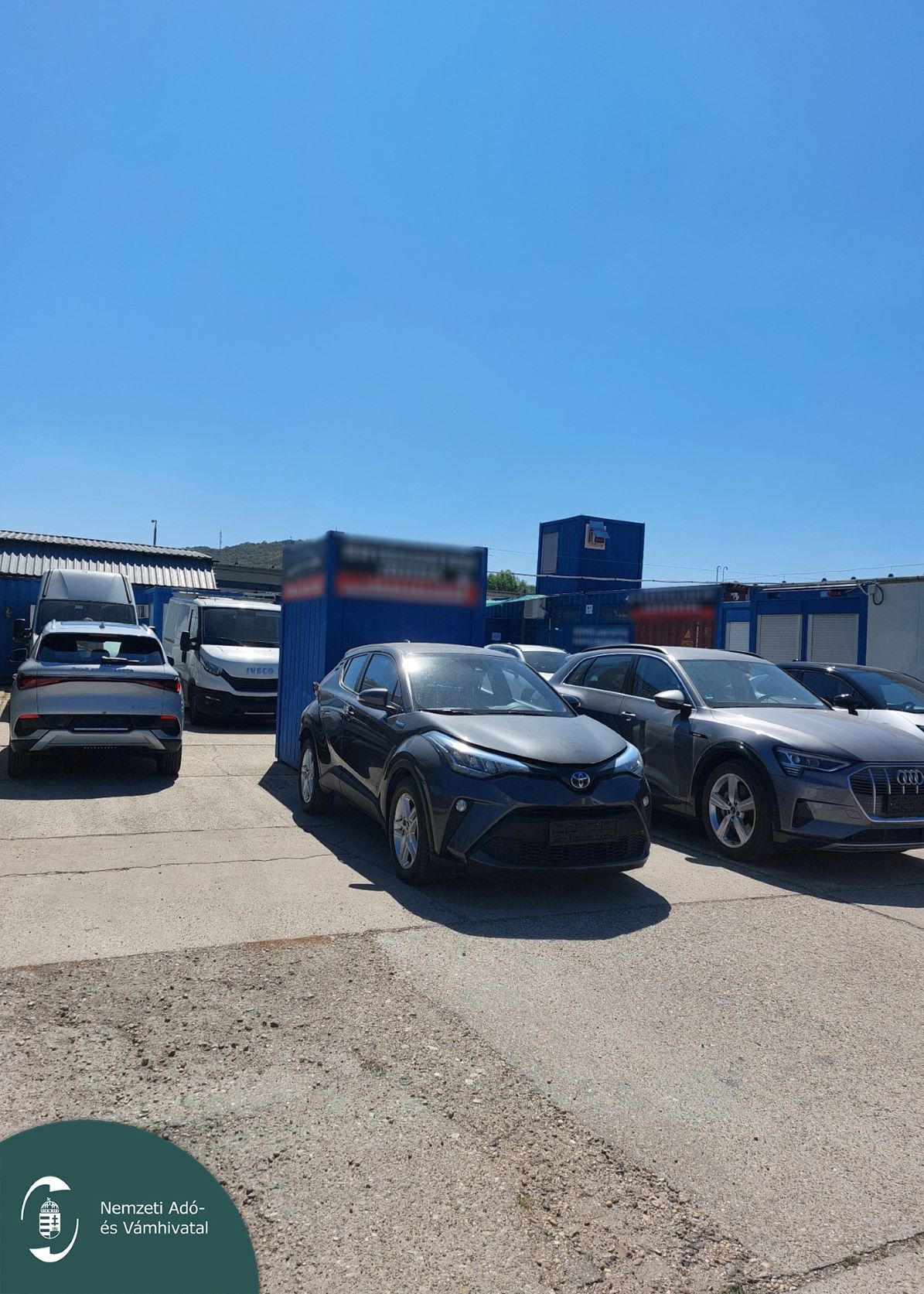 Toyota és Audi személyautók, valamint haszonjárművek a kék konténerépületek előtti autókereskedői udvaron.