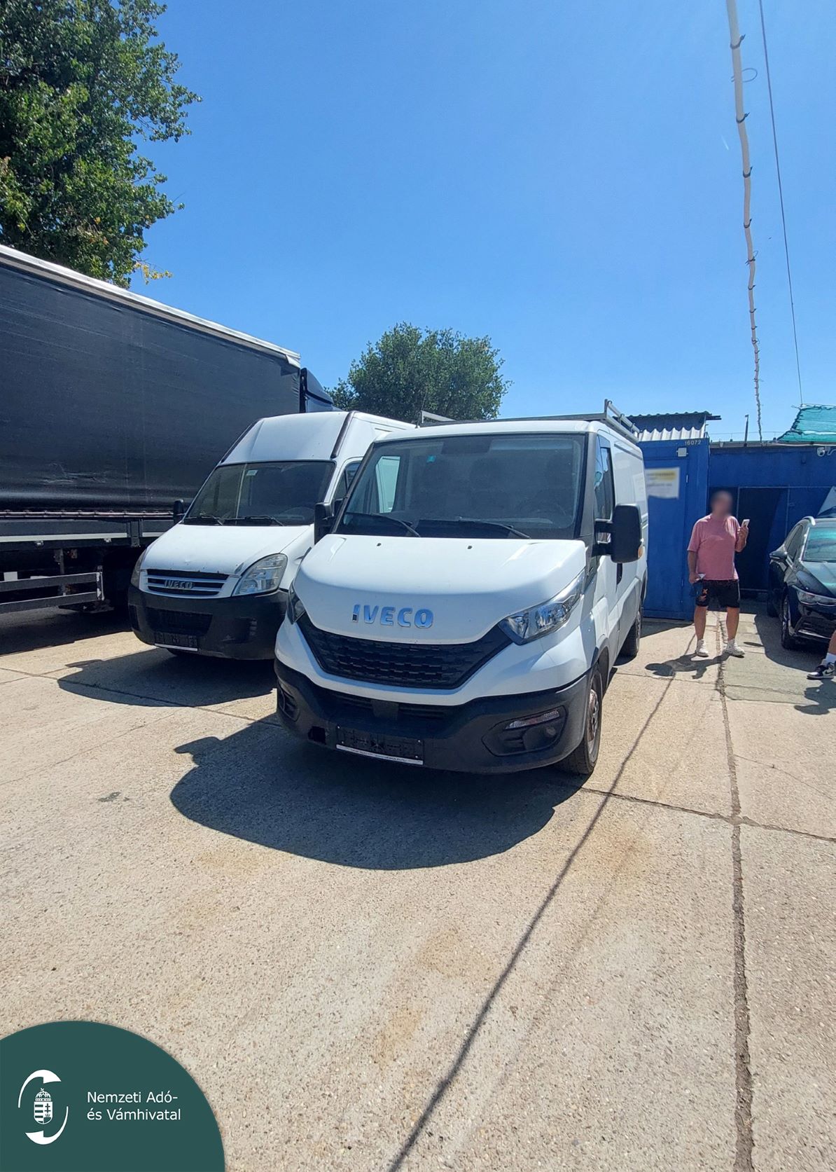 Két fehér Iveco haszonjármű parkol az autókereskedés udvarán, kék konténerépület előtt.