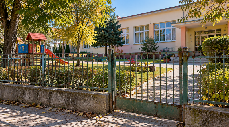 Őszi napsütésben álló óvoda, előtte régi fémkerítéssel és játszótérrel